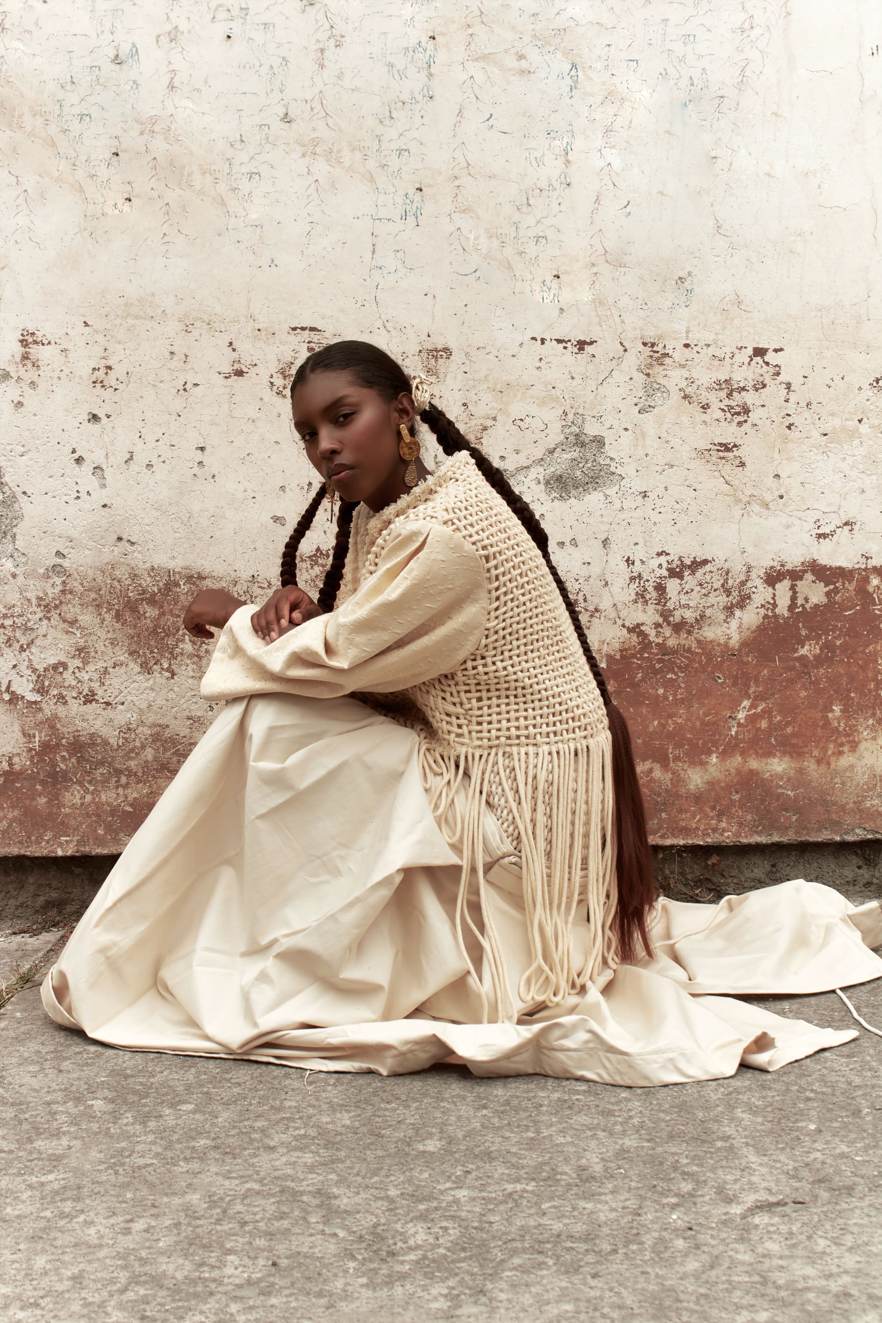 Editorial Coangue: modelo arrodillada con poncho tejido de flecos junto a un muro antiguo.
