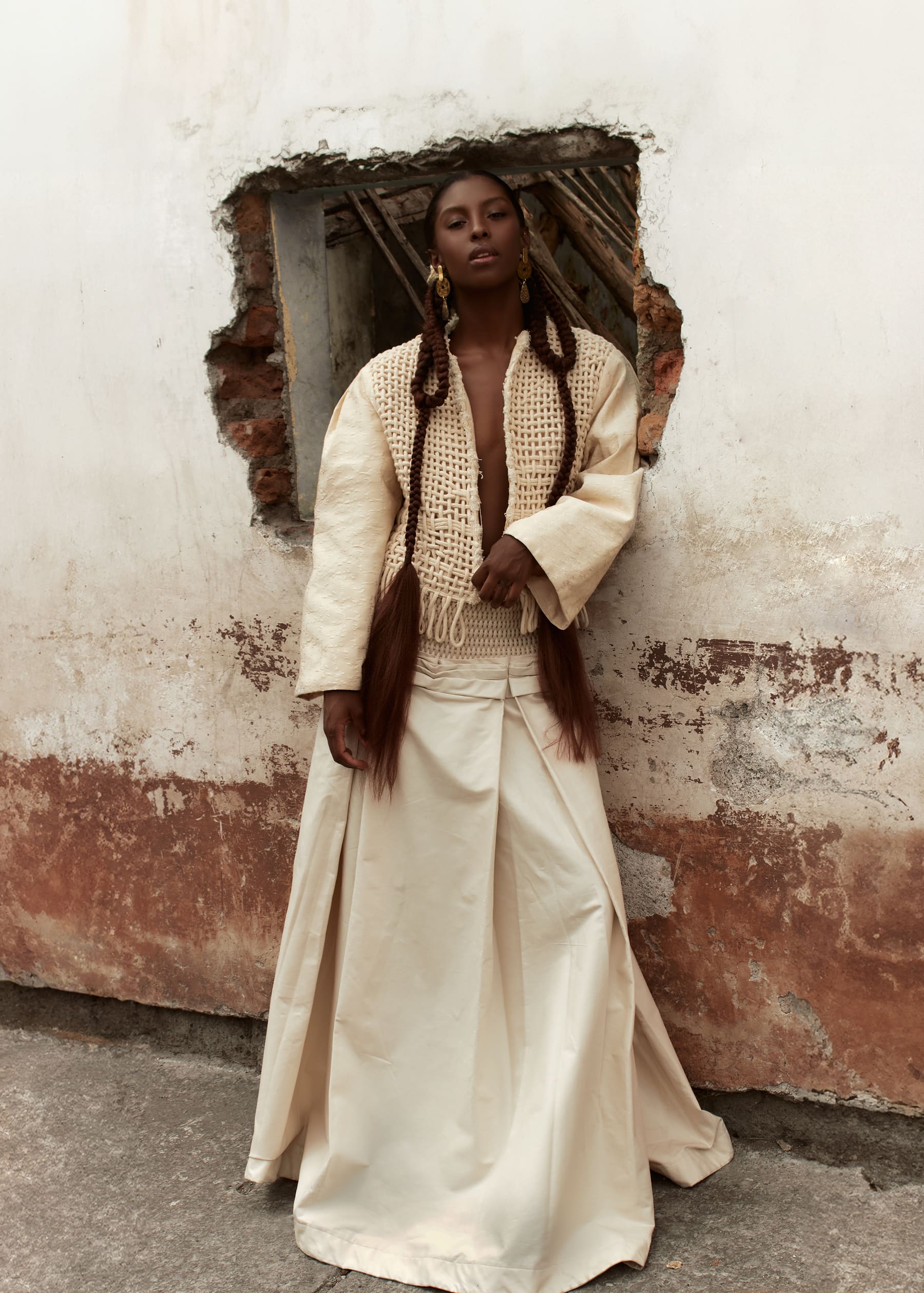 Editorial Coangue: modelo de pie en un vano de pared derruida, con vestido crudo y trenzas largas.