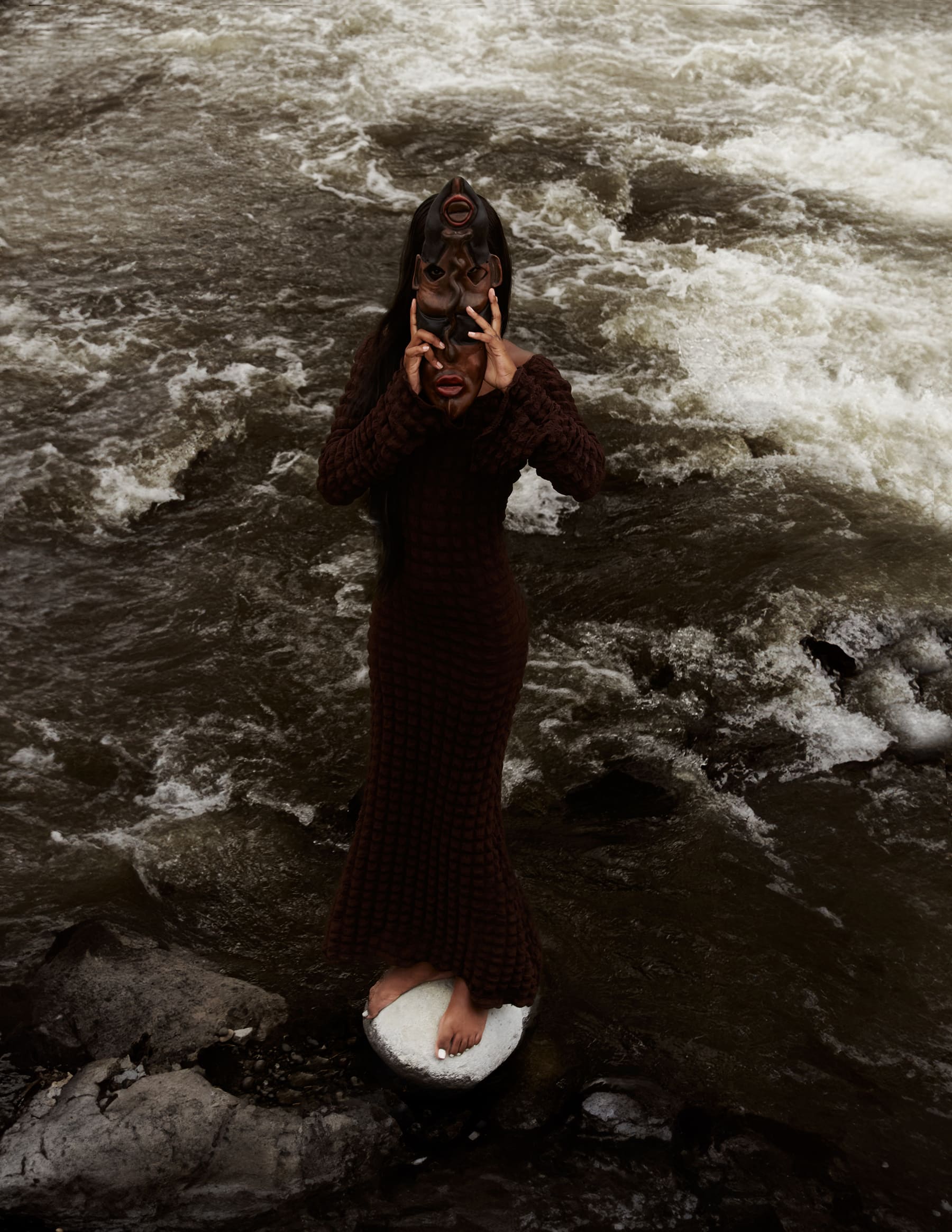 Editorial Coangue: modelo con máscara de madera de pie sobre una piedra en el río.