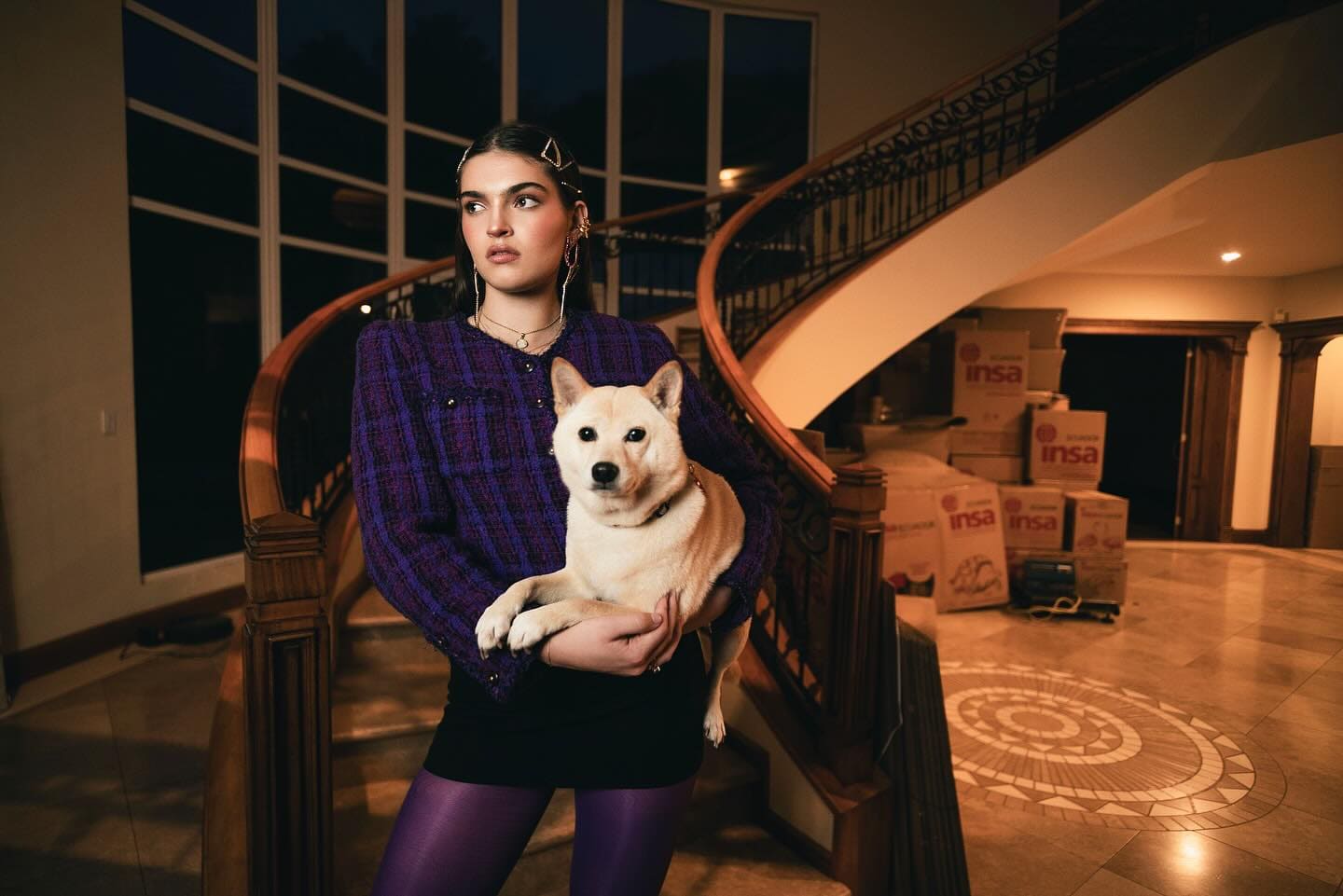 Editorial InStyle Grecia: modelo con blazer morado tweed sosteniendo un perro shiba inu en una escalera curva, cajas de mudanza al fondo.