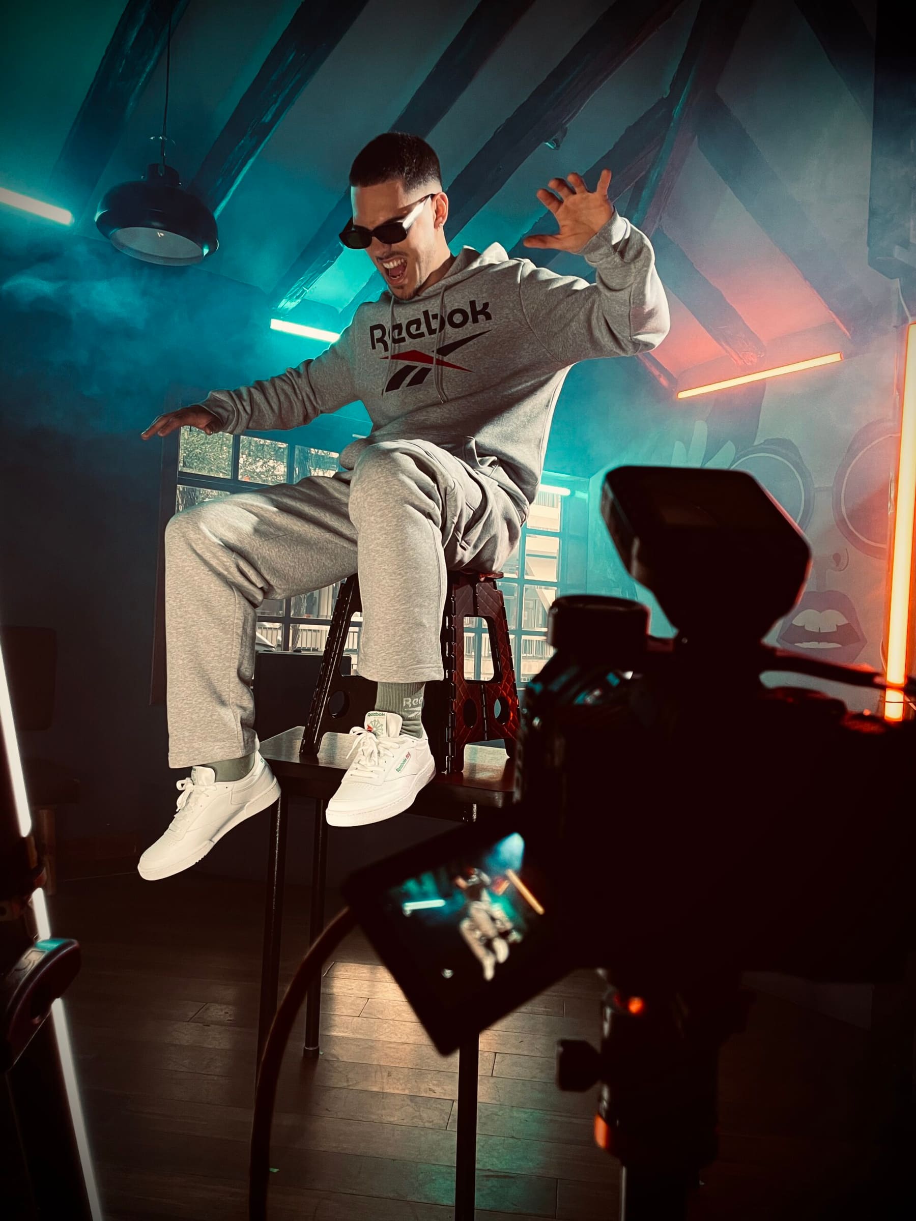 Detrás de cámaras Reebok: modelo agachada junto a equipo de estudio, esfera espejada de fondo.
